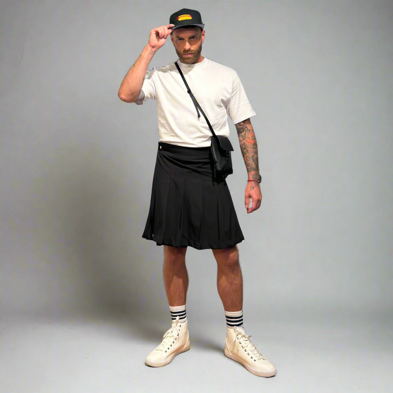 Man wearing a black kilt, white t-shirt, white sneakers, and a black cap on a gray background