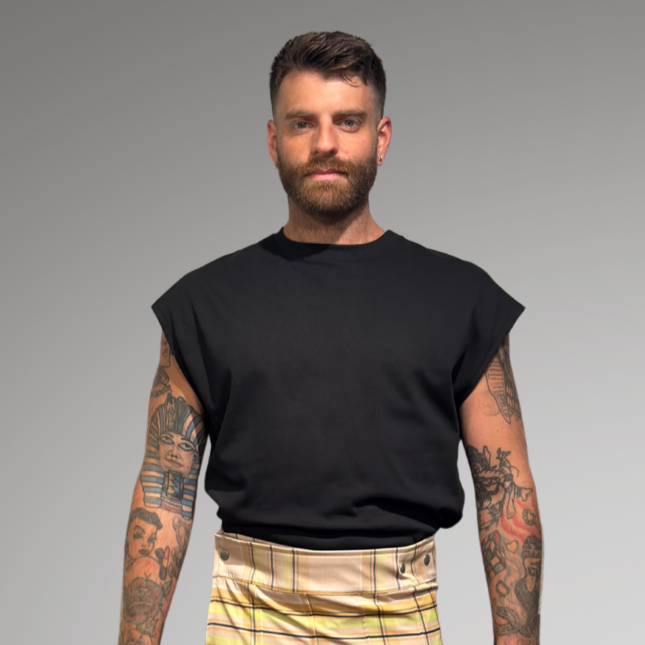 Man wearing a black sleeveless shirt and plaid  kilt against a gray background