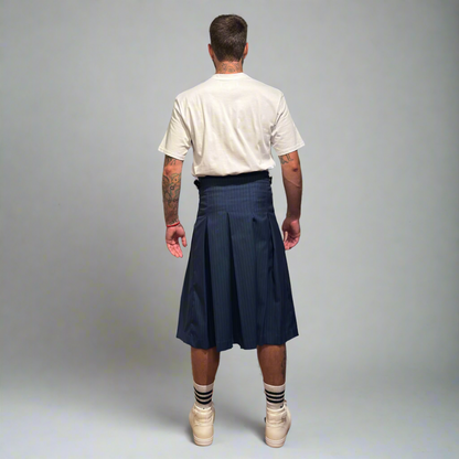 Man wearing a navy pleated kilt with a white t-shirt and white sneakers on a gray background